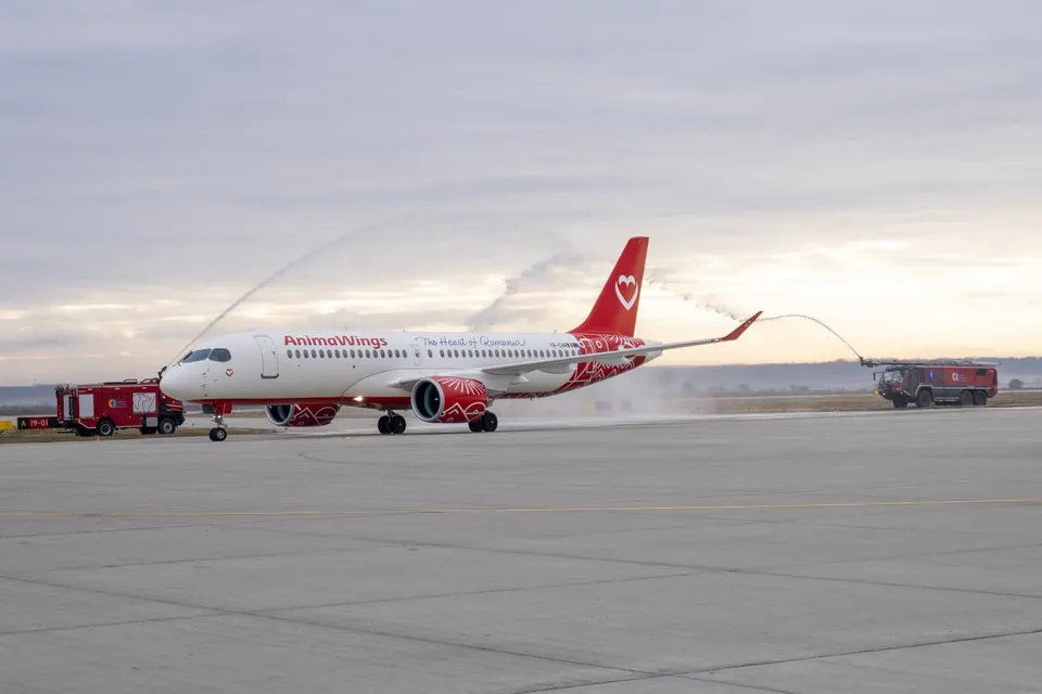Un avion alb cu marcaje roșii este spălat cu jeturi de apă de două mașini de serviciu pe o pistă de beton sub un cer înnorat.