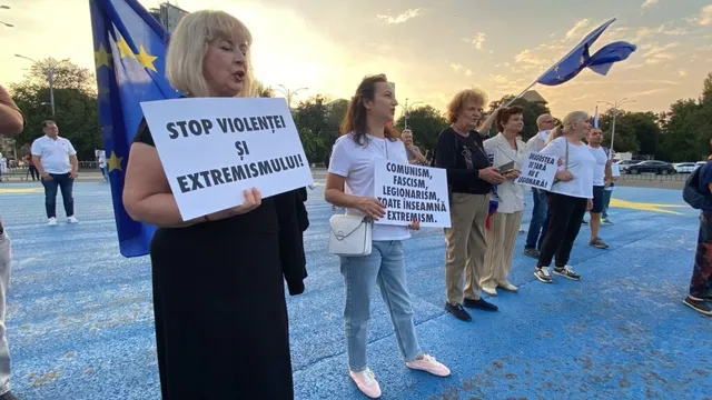 Un grup de persoane stă pe o suprafață albastră și ține diverse panouri albe cu inscripții negre sub un cer de seară.