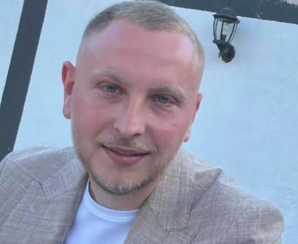 Un bărbat cu păr scurt, purtând un sacou bej și un tricou alb, privește spre cameră în fața unui perete alb.
