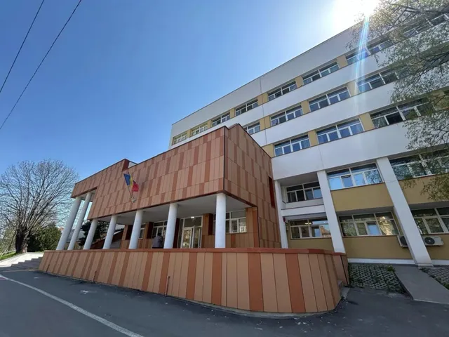 O clădire cu ferestre multiple și panouri maronii dispuse vertical este situată sub un cer senin și însorit lângă un drum asfaltat.