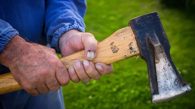 Mâinile unui bărbat ținând un mâner de lemn al unei secure cu cap metalic deasupra unui fundal verde.