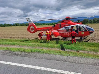 Un elicopter roșu și alb cu inscripția SMURD este aterizat pe un câmp lângă un drum, înconjurat de trei persoane îmbrăcate în uniforme roșii.