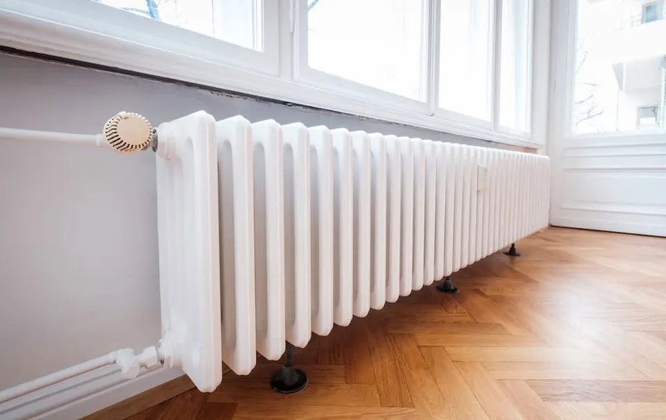Un radiator alb de tip șanț este montat pe un perete lângă o fereastră, deasupra unui podea din lemn.