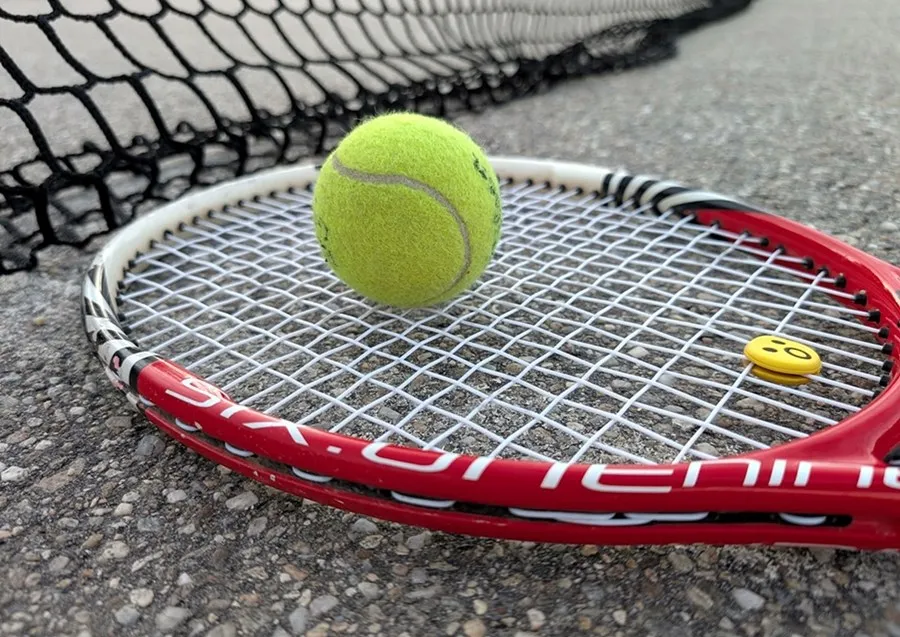 O minge de tenis galbenă este așezată pe coarda unei rachete de tenis roșu și albe, lângă o plasă neagră pe un teren de asfalt.