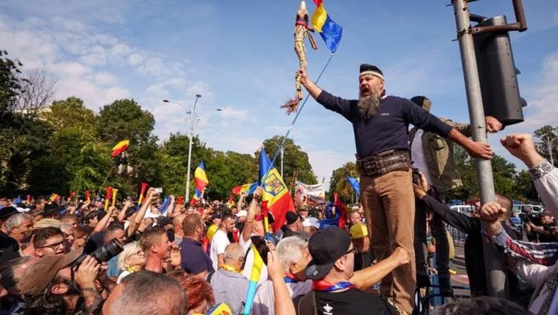 Un bărbat cu barbă, purtând o bandană și un toiag cu steaguri, stă pe un stâlp în mijlocul unei mulțimi de protestatari cu steaguri.