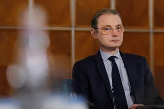 Un bărbat cu ochelari, purtând sacou închis la culoare, cămașă deschisă și cravată, stă așezat în fața unui fundal din panouri de lemn maro.