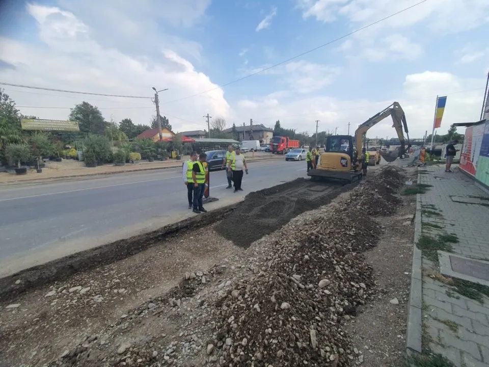 O excavatoare galbenă lucrează pe un drum lângă un grup de persoane purtând veste reflectorizante de siguranță sub un cer înnorat.