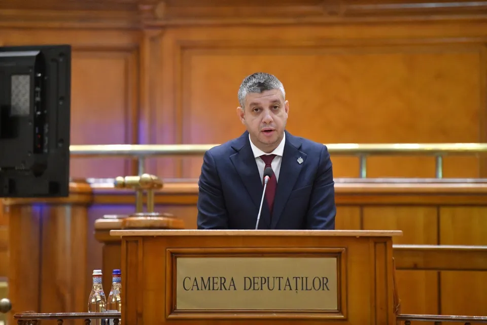 Un bărbat într-un costum închis și cravată roșie stă în spatele unui podium de lemn pe care scrie CAMERA DEPUTAȚILOR.