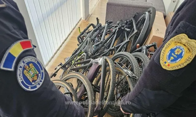 Două persoane în uniformă întunecată stau lângă un grup de biciclete depozitate pe un podea interioară.