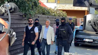 Un grup de bărbați merge pe un drum lângă o clădire, printre care un bărbat cu sacou deschis și mai mulți bărbați în uniformă neagră.