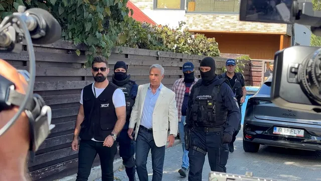 Un grup de bărbați merge pe un drum lângă o clădire, printre care un bărbat cu sacou deschis și mai mulți bărbați în uniformă neagră.