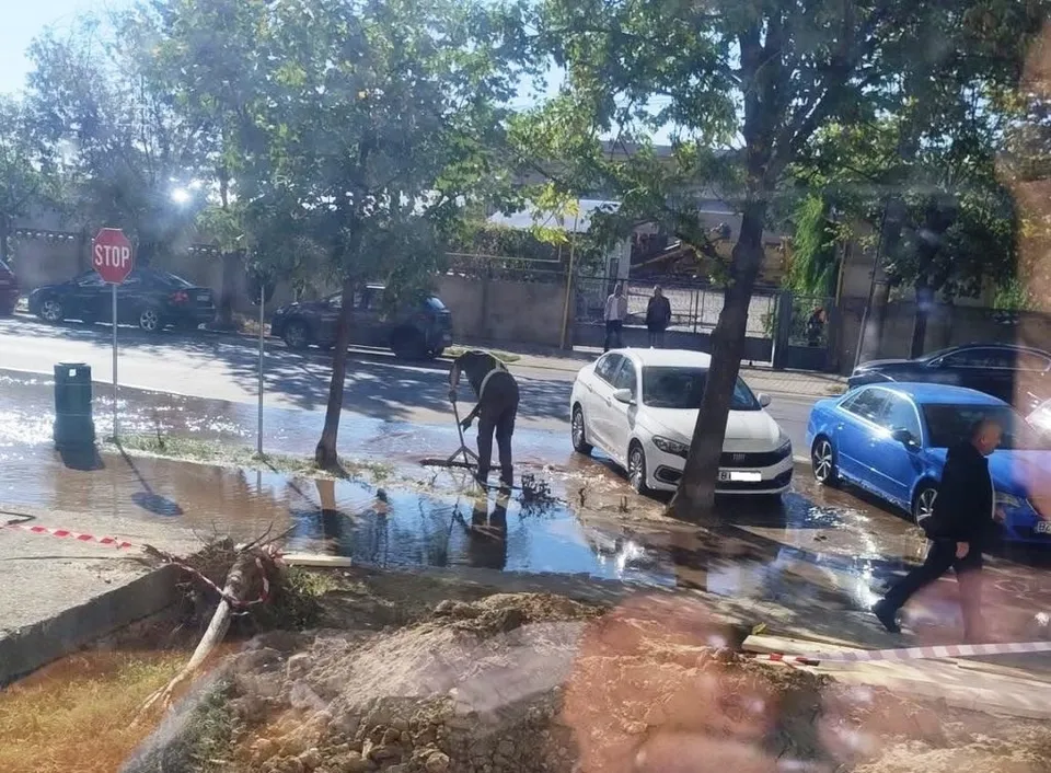 O persoană în haine închise stă lângă o conductă într-o groapă cu apă, pe un drum asfaltat lângă mașini parcate și copaci.