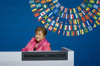 O femeie cu părul scurt și o jachetă roz stă în spatele unui pupitru alb cu inscripția Managing Director.