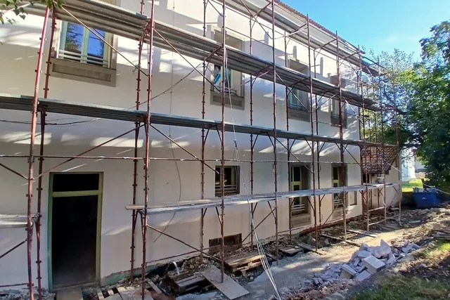 O clădire albă cu ferestre este acoperită de niște schele metalice pe fațadă, având resturi de materiale de construcție pe sol în prim-plan.