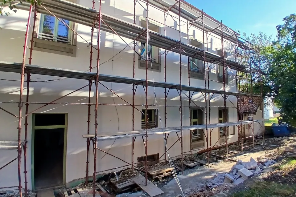 O clădire albă cu ferestre este acoperită de niște schele metalice pe fațadă, având resturi de materiale de construcție pe sol în prim-plan.