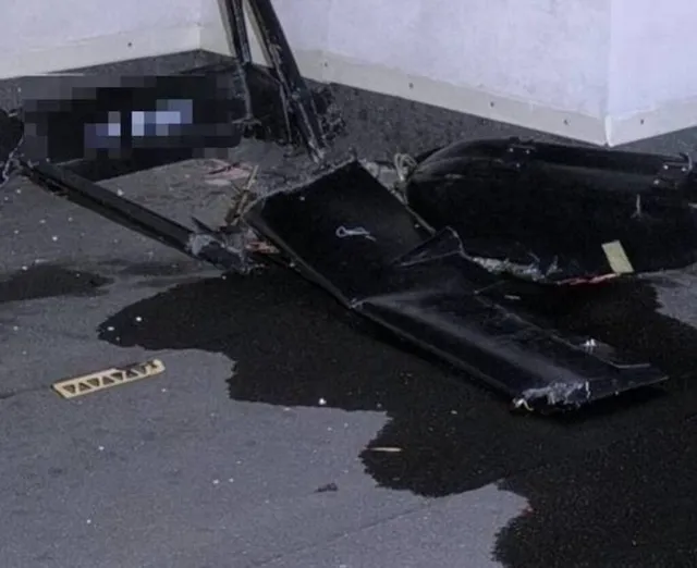 Fragmente negre de un obiect zburător sunt împrăștiate pe o suprafață asfaltată lângă un perete alb.