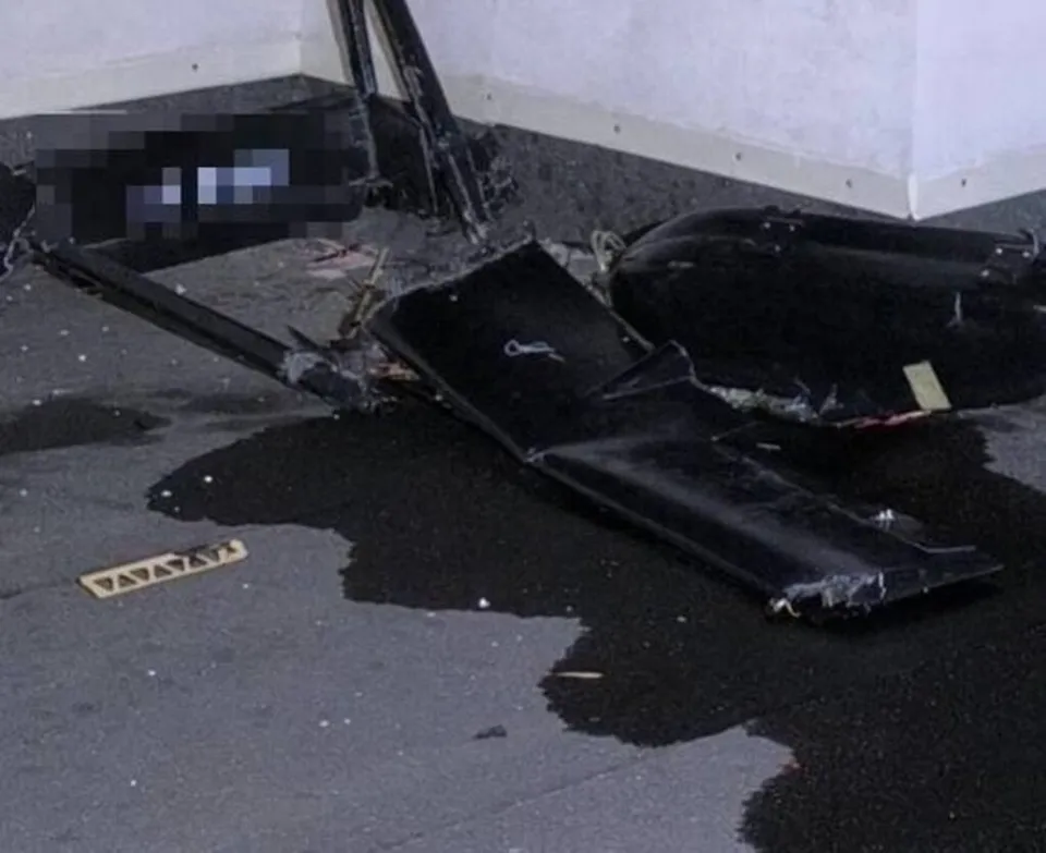 Fragmente negre de un obiect zburător sunt împrăștiate pe o suprafață asfaltată lângă un perete alb.