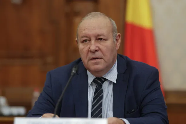 Un bărbat cu părul scurt și costum închis peste o cămașă albastră și cravată în dungi stă la o masă cu un microfon în față.
