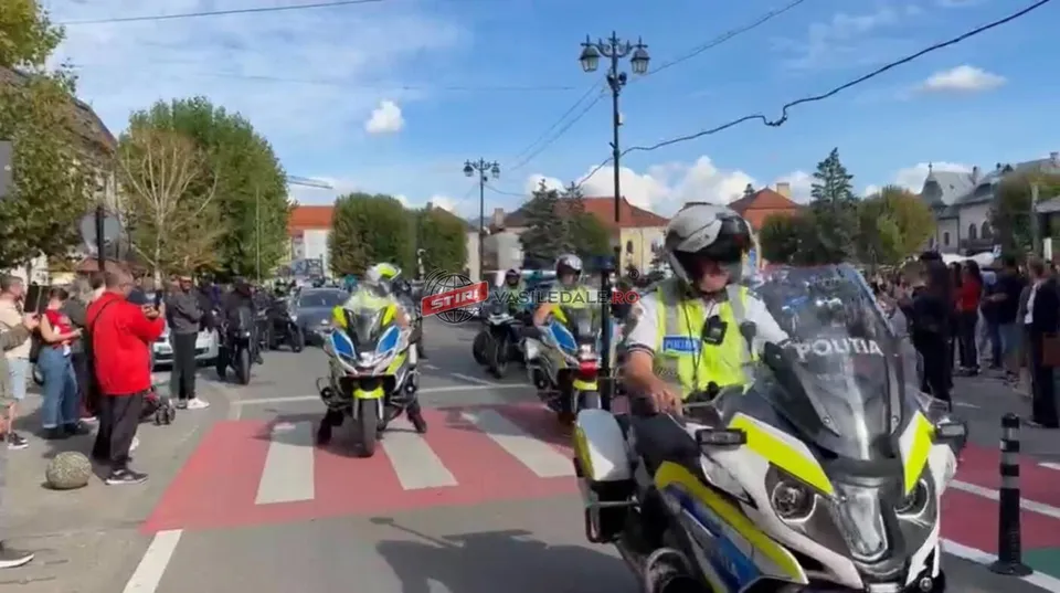 Mai mulți polițiști pe motociclete cu marcajul politia circulă pe un drum asfaltat lângă un grup de oameni stând pe marginea drumului.