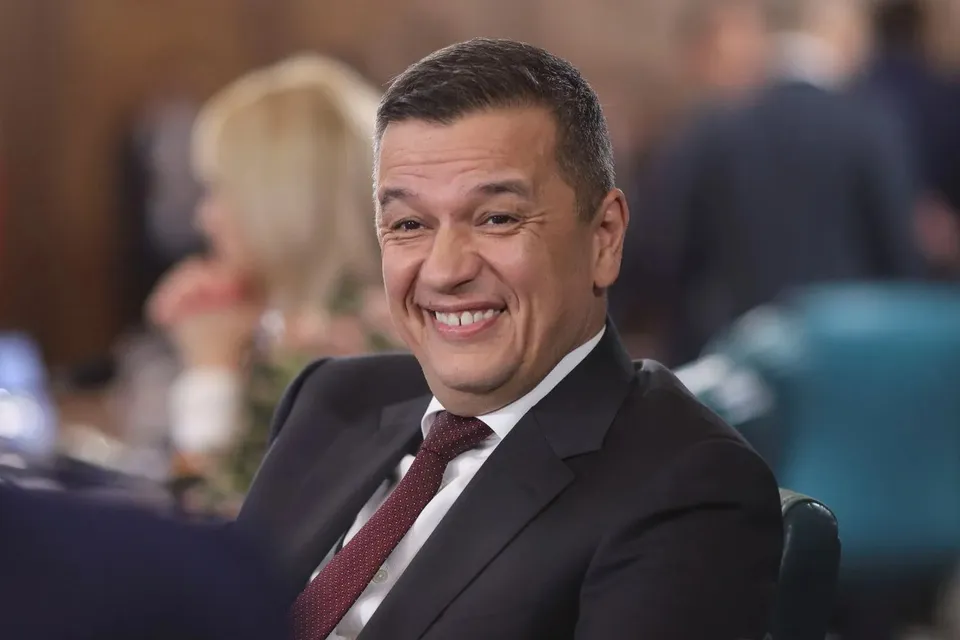 Un bărbat zâmbitor cu păr scurt, purtând un costum închis la culoare, cămașă albă și cravată roșie, stă așezat într-un decor cu persoane în fundal.