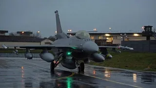 Un avion de luptă gri stă pe o pistă umedă sub un cer înnorat, lângă clădiri și turnul de control.