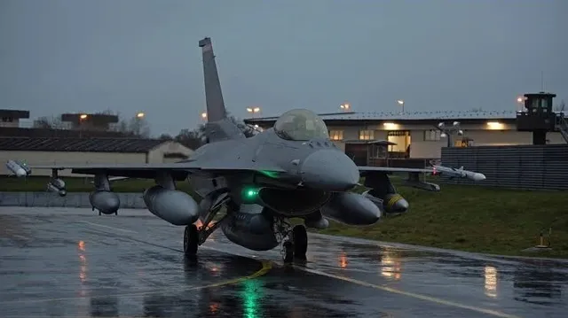 Un avion de luptă gri stă pe o pistă umedă sub un cer înnorat, lângă clădiri și turnul de control.