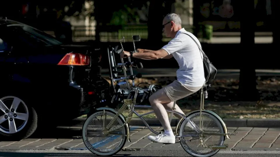 Un bărbat cu ochelari și tricou alb pedalează pe o bicicletă cu trei roți pe lângă o mașină neagră parcată.