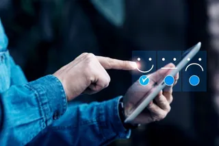 O persoană în cămașă de denim ține o tabletă și atinge ecranul, lângă care sunt afișate trei pictograme de tip emoticon pe un fundal albastru.