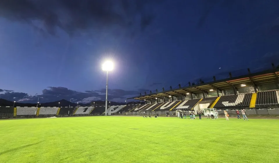Un teren de fotbal verde este iluminat de un stâlp înalt sub un cer de seară, lângă tribunele unui stadion cu scaune galbene și negre.