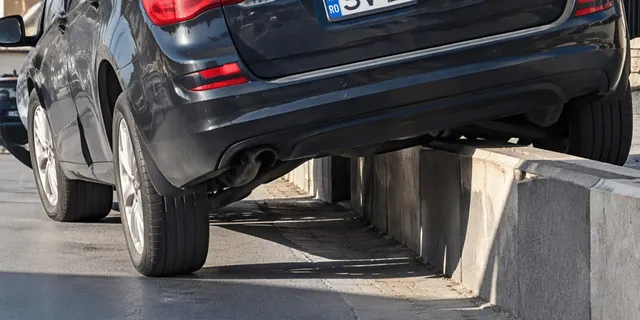 Un autoturism negru este poziționat cu roțile pe asfalt și pe un bord curb, fiind înclinat pe partea dreaptă.