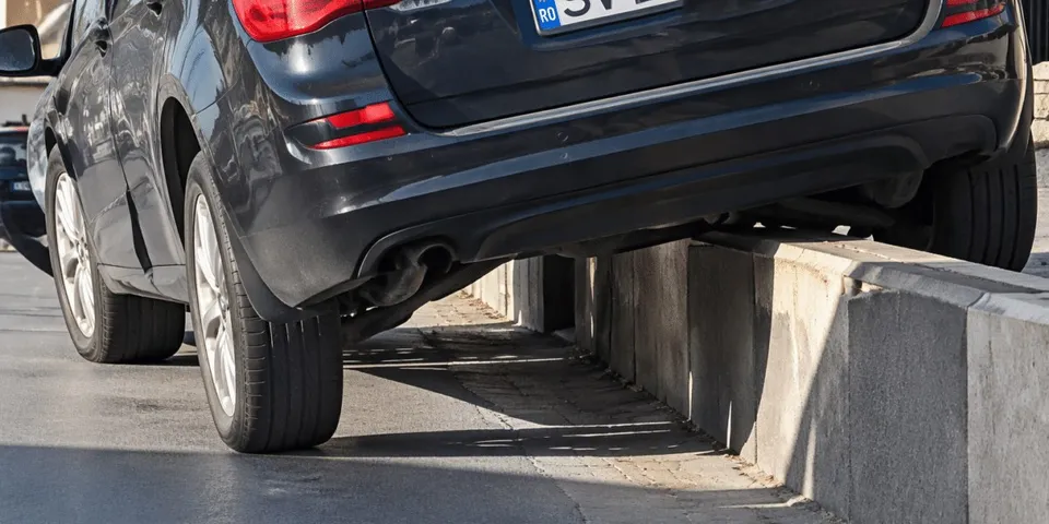 Un autoturism negru este poziționat cu roțile pe asfalt și pe un bord curb, fiind înclinat pe partea dreaptă.