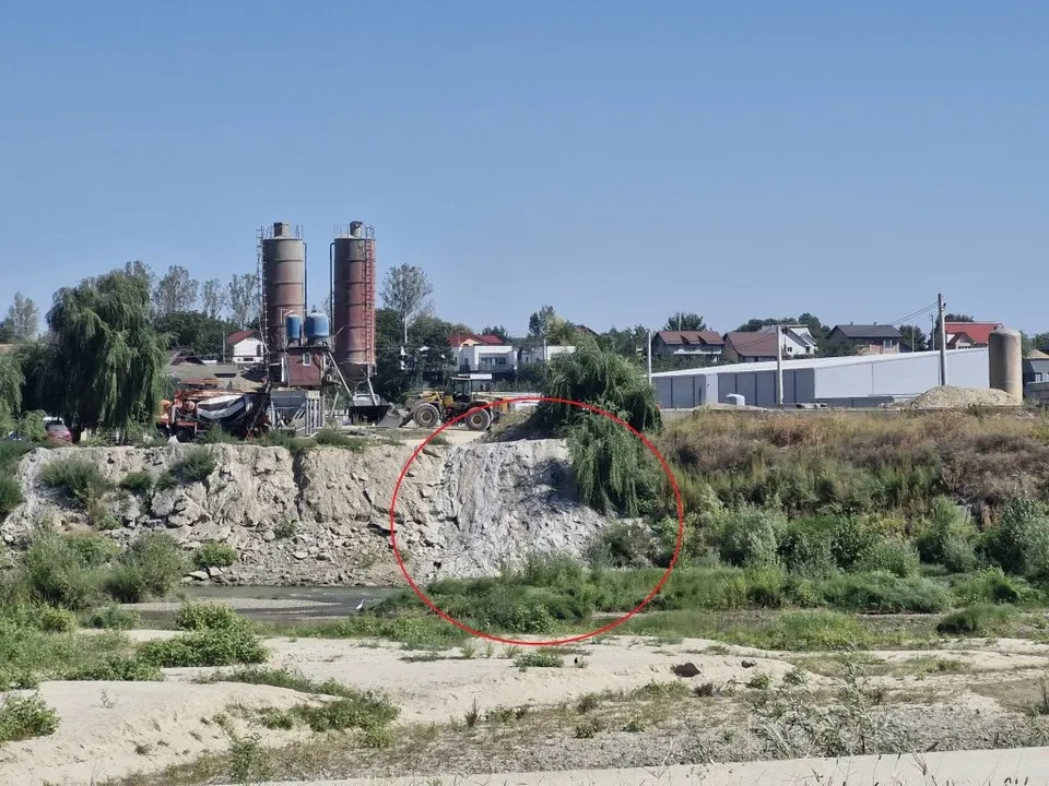 Două silozuri înalte se ridică lângă câteva clădiri, în timp ce un teren nisipos și vegetație joasă ocupă prim-planul sub un cer senin.