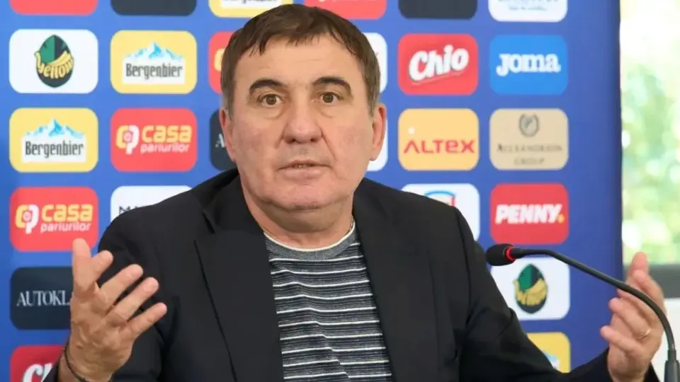 Un bărbat cu părul scurt și brunet poartă un sacou negru peste un tricou gri cu dungi orizontale, stând în fața unui fundal colorat cu diverse logo-uri.