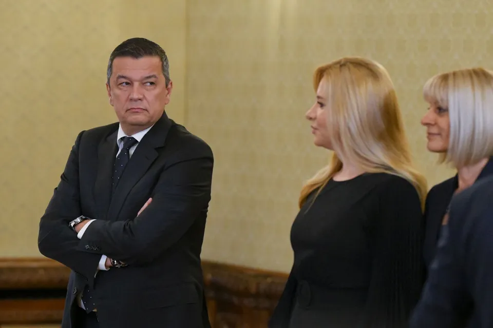 Un bărbat într-un costum negru stă cu brațele încrucișate, privind spre două femei care stau în profil în fața lui.