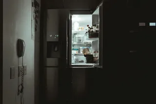 Un frigider deschis într-o încăpere întunecată scoate lumină, afișând diverse alimente și recipiente pe rafturile interioare.
