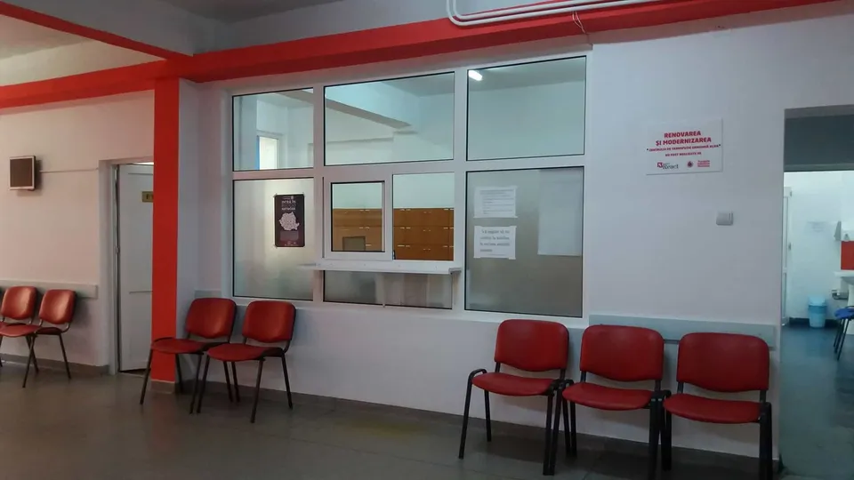 O cameră cu pereți albi și detalii roșii conține mai multe scaune roșii aliniate și o fereastră de serviciu cu un ghișeu.