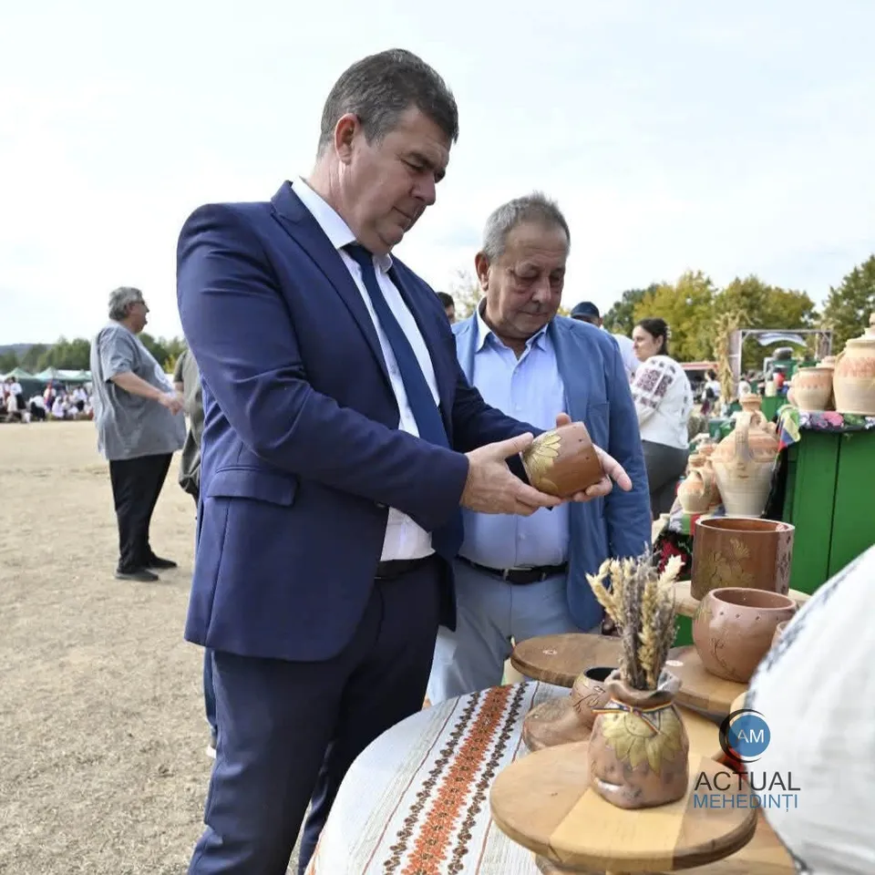 Un bărbat într-un costum albastru ține un vas de lut maro, în timp ce un alt bărbat îl privește de lângă el.