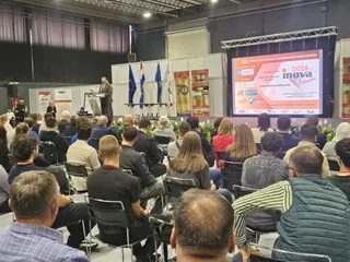 O mulțime de oameni stă pe scaune în fața unui podium unde un bărbat vorbește lângă un ecran mare și niște steaguri.