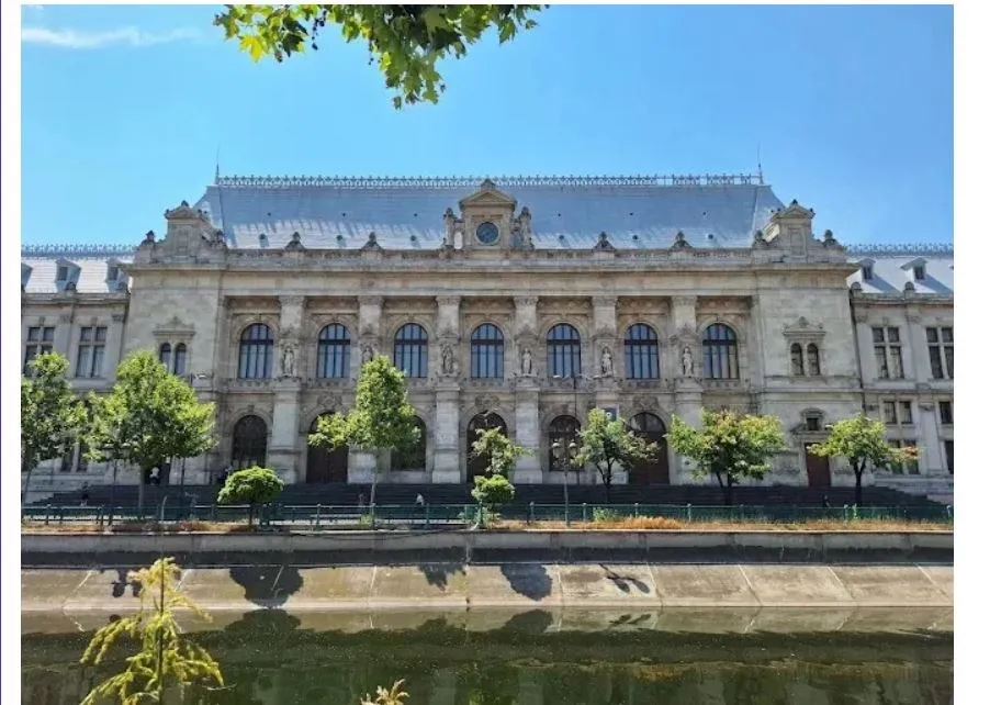 O clădire monumentală de piatră cu ferestre înalte și arcade este situată în spatele unor copaci verzi și a unui gard metalic.