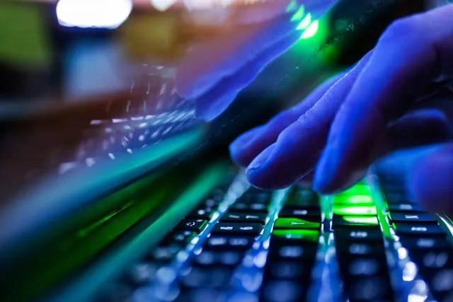 Mâini deasupra unui tastatură iluminată de lumini verzi și albastre într-un mediu întunecat.