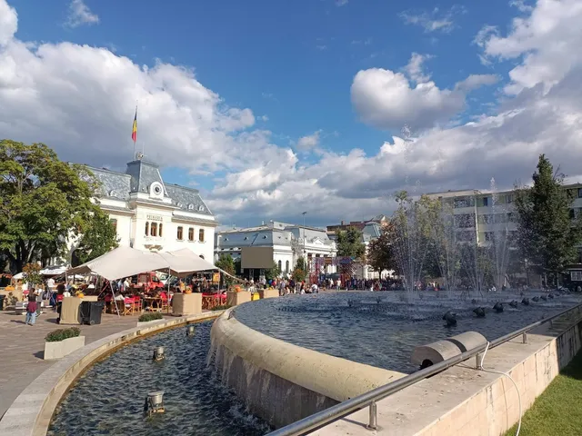 O fântână cu jeturi de apă se află într-o piață publică, lângă mese cu umbrele de grădină și clădiri în fundal sub un cer înnorat.