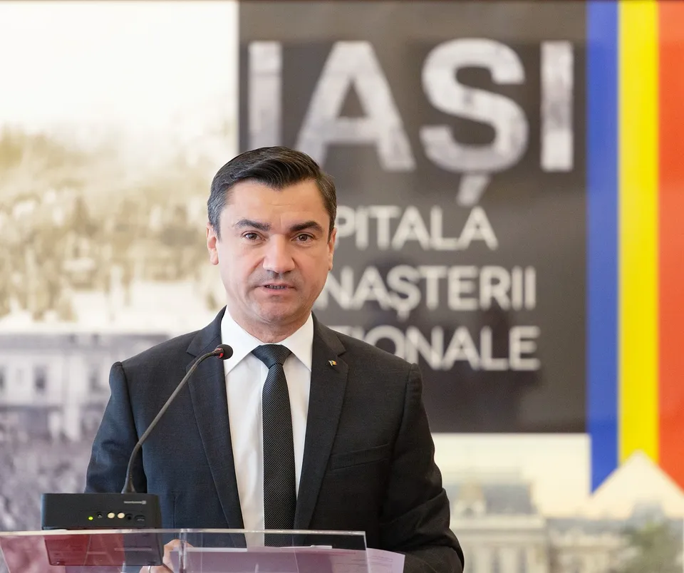 Un bărbat cu părul scurt, purtând costum negru, cămașă albă și cravată neagră, stă în fața unui microfon în fața unui panou cu textul IAȘI.