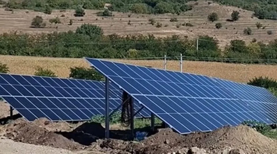 Mai multe panouri solare albastre sunt montate pe structuri metalice într-un teren cu dealuri și vegetație în fundal.