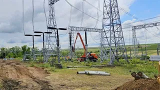 O șantier de construcție cu un macara portocalie, stâlpi de tensiune înalți și un șanț de pământ într-un peisaj rural.