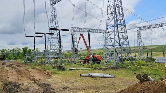 O șantier de construcție cu un macara portocalie, stâlpi de tensiune înalți și un șanț de pământ într-un peisaj rural.