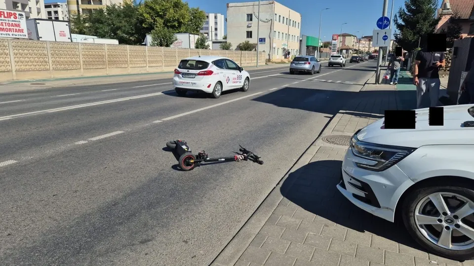 O trotinetă electrică neagră este întinsă pe asfaltul unei străzi lângă un autoturism alb parcat pe marginea trotuarului.