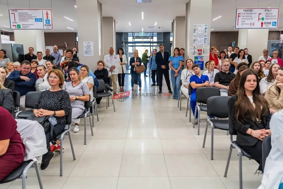 Oamenii stau așezați pe scaune negre într-un hol cu podea deschisă, orientați spre un spațiu central de circulație.