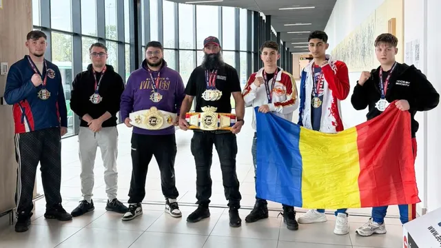 Șapte bărbați stau în rând într-o încăpere luminoasă, purtând medalii și ținând două centuri de campion și un steag tricolor.