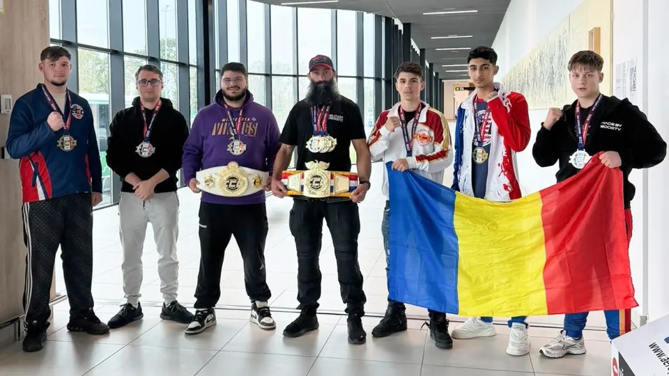 Șapte bărbați stau în rând într-o încăpere luminoasă, purtând medalii și ținând două centuri de campion și un steag tricolor.
