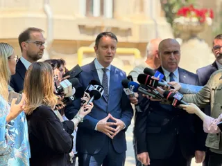 Un bărbat într-un costum albastru și cravată stă în centru, înconjurat de microfoane și persoane care îi adresează întrebări.